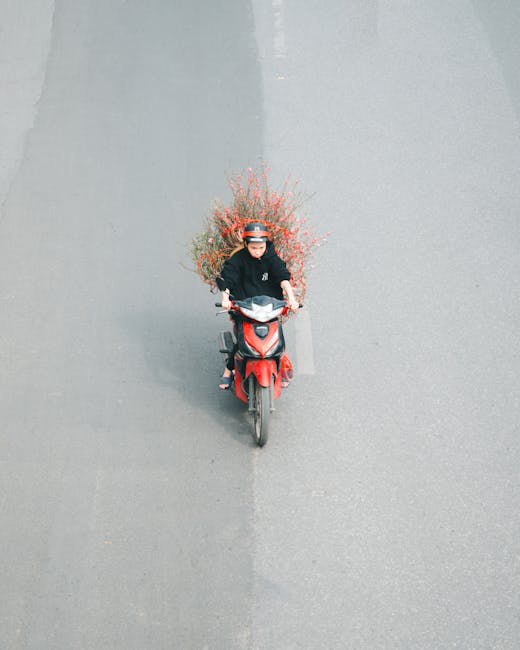 A delivery person in a black jacket and helmet rides a red motorbike on a grey road, carrying a large bouquet of fresh flowers extending high above their head. The bouquet features pink, orange, and g