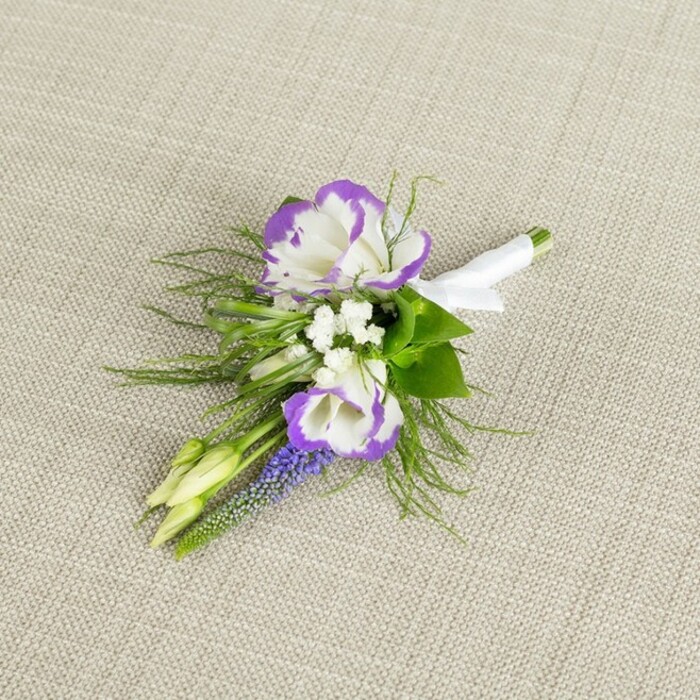 Set against a softly textured, light beige textile that looks like fine linen, this elegant boutonniere is captured in a close-up that reveals the care of an experienced florist. The eye is immediately drawn to two white lisianthus blooms at the centre, their petals gently unfurling in layered ruffles, each edge finely traced with a rich, royal purple that creates a dramatic yet refined contrast. These central flowers are surrounded by tiny, crisp white filler blossoms, reminiscent of baby's breath, which lend a delicate, cloud-like fullness and a sense of lightness around the more defined shapes of the lisianthus. To one side, a slim spike of muscari stands upright, made of tiny, tightly packed, bell-shaped florets in a deep purple-blue that deepens the palette and adds a graceful vertical accent, much like a slender tower rising above a Queensbury garden bed. Fine, feathery greenery, similar to asparagus fern, spills out in soft sprays, its thin, threadlike fronds catching the light and contributing texture and movement, echoing the gentle sway of foliage on breezy days near Queensbury's tree-lined streets. Two closed, pale green buds nestle within the greens, their smooth, tapered forms suggesting flowers that will open in time, a subtle nod to new chapters starting. The stems of all these elements are carefully aligned and tightly bound, then wrapped in a clean band of white satin ribbon that is smoothed flat and finished with a neat, understated bow, clearly prepared for easy pinning to a suit or jacket. The overall feeling is one of timeless, understated luxury, as if this boutonniere is ready for a groom or groomsman attending a classic ceremony in one of Queensbury's local venues or a smart celebration just a short drive from the town centre.