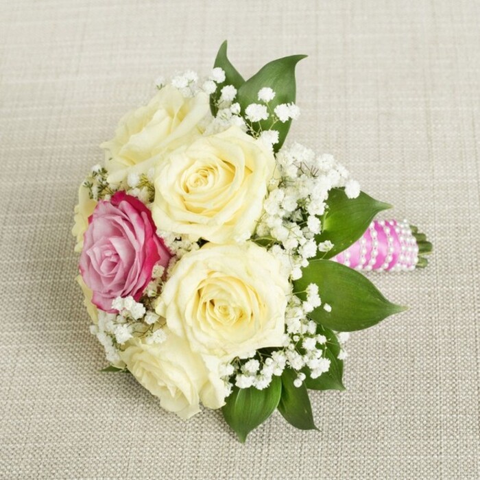 Viewed from above, this compact bridal bouquet in Queensbury is a study in exquisite detail and romantic storytelling. Its creamy, pale yellow roses-five or six in full, dew-kissed bloom-are clustered together, their unspoiled petals freshly unfurled, suggesting the hopeful promise of a wedding day. Just off-center, a dazzling magenta-pink rose draws the eye and warms the heart, its lively hue reminiscent of the blossoms often found at Canons Park on a bright spring morning. Fine sprays of feathery white baby's breath are artfully woven throughout, lending a sense of airiness and traditional bridal elegance, accentuated by the deep green leaves at the bouquet's lush edges. The stems, extending neatly to the right, are enveloped in a satin ribbon of rich, fuchsia-pink-its sheen capturing soft natural light and evoking luxury. Adorned with interlacing strands of luminous pearls wound in perfect spirals, the handle sparkles subtly, much like the joyous laughter carried from nearby Queensbury celebrations. Set against a backdrop of pale, woven fabric, the bouquet stands as a testament to local florist craftsmanship. Tailored for weddings, anniversaries, or meaningful events, this bouquet brings together the spirit of Queensbury, expert floral design, and timeless celebration.