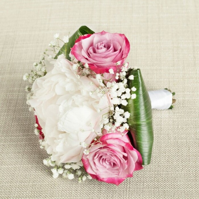 Picture a stunning, locally inspired bouquet from the heart of Queensbury, arranged on a pale, textured linen that grounds the composition in understated luxury. Dominating the arrangement are lush, full white peonies, their ruffled, cloud-like petals spilling outwards in waves that catch the gentle light and speak of pure celebration. Two striking pink roses, their nuclei tightly curled and their outer petals glowing from hot fuchsia to delicate blush, anchor the bouquet with timeless beauty. Effortlessly interspersed are fragrant sprigs of baby's breath-each tiny blossom reminiscent of spring mornings near Kingsbury Road-offering an ethereal, airy contrast. The bouquet is expertly shaped with broad, deep green leaves, likely fresh aspidistra or ti foliage, folded to cradle the arrangement and impart structure and depth. Around the base, a pristine white satin ribbon envelops the stems as a classic finishing touch, its soft sheen adding to the bouquet's polished appeal. The balanced lighting reveals every nuance: the texture of peony petals, the gentle twinkle of gypsophila, the subtle gloss of each green leaf. Ideal as a bridesmaid's bouquet for local weddings or as a statement gift for milestone events celebrated in Queensbury's proud community, this design is a beautiful testament to local craftsmanship and heartfelt artistry.