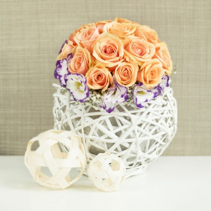On a clean white surface that could easily be a dining table in a Queensbury home or a dressed table at a local wedding venue, a meticulously crafted floral centerpiece takes focus. At the top sits a dense, spherical mass of roses in shades of peach and soft apricot, each bloom fully open and arranged so closely that the petals blend into one continuous, velvety dome. The roses have a gentle, romantic glow, their warm tones conveying tenderness and celebration. Encircling the base of this rose globe is a ring of white lisianthus blooms, their ruffled petals finely outlined in bold violet-purple, as if each flower has been brushed with ink around the edges. Delicate sprigs of white gypsophila peek through, softening the contrast and lending the arrangement a weightless, airy feel. The flowers are gathered into a distinctive white vessel made from interwoven rattan or wicker that bulges outward in a rounded, bulb-like shape. The open lattice structure of the container allows light to permeate, highlighting shadows between the strands and giving the whole piece a sculptural quality. In front of the vase, two pale, woven spheres lie on the same surface, fashioned from narrow, natural-toned wooden strips that loop and criss-cross to create open, spherical forms; one is larger, the other smaller, like minimalist companions to the main design. The backdrop is a soft, beige fabric with a gentle texture, reminiscent of linen or burlap, which acts as a quiet canvas so the floral colours can sing. The composition suggests understated luxury and timeless romance, ideal for wedding table arrangements, anniversary dinners, or sophisticated floral styling for Queensbury events where elegance and comfort meet.