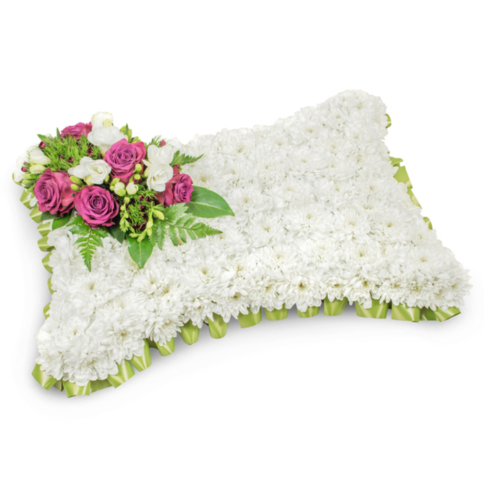 Presented like a small, serene cushion of flowers, this rectangular pillow memorial is densely covered with hundreds of immaculate white chrysanthemums whose layered petals form a smooth, plush surface. The uniform white field conveys calm and purity, its texture soft and slightly fragrant with green, floral notes that remind one of quiet mornings. On the top-left corner a carefully arranged accent of deep fuchsia or magenta roses offers a poignant pop of colour; their open, velvety faces are softened by clusters of tiny white freesias and buds that add lightness and a gentle scent. Slender fern fronds and glossy leafy greens tuck around the accent, framing it and giving depth and movement to the arrangement. A narrow pleated border of pale green satin ribbon encircles the pillow, discreetly peeking out and giving the piece a refined edge that feels both respectful and thoughtfully finished. Against a crisp white background the pillow reads with clarity and dignity - a traditional sympathy design meant to convey peace, rest, and lasting remembrance. As a Queensbury-based florist familiar with local funeral directors and church services near the high street and surrounding suburbs, I would recommend this cushion for services where families seek a timeless expression of condolence: understated, fragrant, and composed with quiet care to cradle memory and offer solace to those who gather.