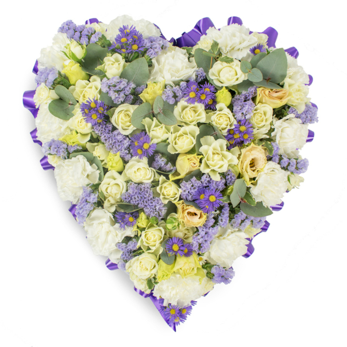Photographed on a crisp white surface with gentle studio illumination, this heart-shaped floral tribute is densely arranged to read as a single, consoling statement. The composition is anchored by numerous creamy roses - pale yellow and soft cream tones that move from tight buds to open faces - creating a plush, layered texture that feels both refined and tender. Ruffled white carnations are interspersed to lend volume and a time-honoured softness, their frilly petals catching the light and forming comforting tonal highlights. Delicate sprays of lavender-coloured statice and clusters of small purple asters with vivid yellow centers are woven through the heart, their tiny, starry shapes offering playful punctuation and depth against the softer blooms. Blue-green eucalyptus leaves are set throughout, their rounded forms and cool, silvery tint giving the design organic rhythm and a calming, aromatic suggestion. The entire heart is bordered by a neat, looped purple satin ribbon that traces the silhouette and frames the memorial with an elegant finish. As a piece intended for sympathy and remembrance, it communicates enduring affection, peace, and respect; one can easily imagine it placed at a funeral service near Queensbury High Street, at a chapel close to Kingsbury Road, or alongside a graveside plaque. The skilled arrangement reflects florist craftsmanship and a careful selection of textures and hues to console those in mourning.