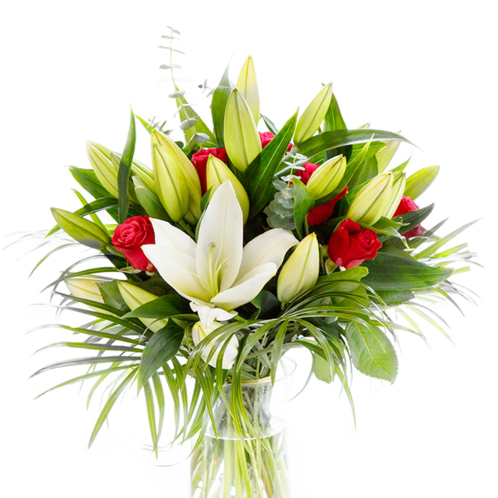 A striking, professionally styled bouquet photographed against a bright white field, where every hue and texture is clearly defined. The arrangement centres on a single, magnificent white oriental lily in full bloom: its pristine, slightly recurved petals form a luminous cup around subtle pale yellow stamens and a delicate green pistil. Radiating outwards are many lily buds, from lime-green to soft yellow, some tightly closed, others in the first breaths of opening, creating a layered rhythm across the bouquet. Interspersed among the lilies are clusters of small, richly colored red roses and tight rosebuds, their velvety petals still mostly rolled inward and delivering concentrated notes of passion within the more tranquil whites and greens. The floristry is supported by diverse foliage: deep, forest-green broad leaves lend structure; longer, feathered palm-like fronds spill gently to one side, adding graceful movement; and slim stems of silvery eucalyptus with rounded leaves introduce cool tonality and a delicate texture. The blooms are set in a classic clear glass vase which reveals tidy, pale green stems submerged in pristine water - a simple vessel that emphasises the flowers' natural beauty. The image evokes a bouquet both refined and warm, suitable as a romantic gesture, a celebratory centrepiece, or a composed sympathy arrangement. I imagine this piece placed on a windowsill near Queensbury High Street or shared at an intimate gathering in the Kingsbury and Harrow border, its scent a harmonious blend of lily's clean powder and the rose's deeper sweetness.