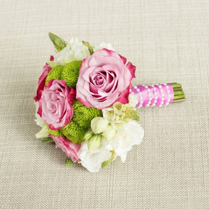 A carefully crafted hand-tied bouquet is shown resting on a neutral, light tan fabric with a visible weave, much like a linen cloth laid out for a small celebration in a Queensbury home. The arrangement is compact and refined, its colours centred on pink, lime green, and white, evoking a fresh springtime stroll through local gardens. Two larger pink roses and a third slightly smaller bloom take centre stage, their petals layered in tight spirals. The colour shifts gently from a soft, romantic blush at the inner whorl to richer fuchsia and magenta along the outer edges, creating depth and a sense of movement within each flower. Around these roses, perfectly rounded lime green button chrysanthemums-those cheerful, pom-pom-like Santini mums-nestle closely, their dense, small petals forming textured spheres that pop against the softer rose petals. Filling in the gaps, creamy white and ivory blossoms, possibly lisianthus or freesia, unfold in delicate, multi-petaled blooms, bringing an airy, almost cloud-like lightness to the composition. Scattered among them are tiny, star-shaped white flowers with golden centres that suggest wax flower or stephanotis, lending intricate detailing a bride might admire up close. The stems are clipped short and bound into a neat posy handle. Vivid fuchsia satin ribbon is wrapped smoothly around the stems, its sheen catching the gentle, diffused light, while a slender strand of white faux pearls spirals over the ribbon in a criss-cross pattern, like a piece of bridal jewellery. A few millimetres of fresh green stem extend beyond the ribbon, cleanly cut. The scene is evenly lit, with no harsh shadow, allowing every texture-from velvety petals to crisp mums and glossy ribbon-to stand out. Created in the style of bespoke designs from Flower Delivery Queensbury, this bouquet feels ideal for a bridesmaid, prom date, or intimate celebration dinner in and around Queensbury.