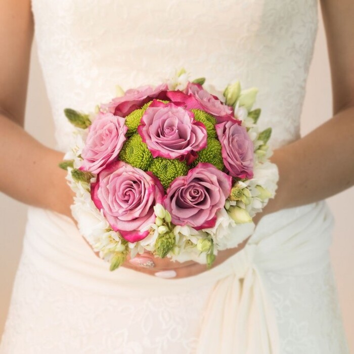 Centrally framed and bathed in gentle light, this image captures a bride in Queensbury holding a meticulously arranged, rounded bridal bouquet. Lustrous roses, with gradients from soft mauve in their core to intense fuchsia along the curled edges, dominate the ensemble; their voluptuous forms are a symbol of enduring romance. Encircling the roses, a ring of lively lime-green button chrysanthemums adds a bright, textured contrast, reminiscent of the green expanses surrounding Church Lane or Queensbury Park. The outer rim dazzles with a halo of creamy white spray roses or perhaps sweetly scented freesia, each tiny blossom accentuated by sprigs of fresh green buds and leaves that enhance the bouquet's depth. The bride's off-white gown-a masterwork of floral lace across the bodice and gracefully draped fabric at the hip-serves as a tranquil canvas, highlighting the artistry and seasonal freshness of the flowers. Her manicured hands, finished in pastel pink with a touch of sparkle on one finger, cup the bouquet gently, radiating a sense of calm anticipation. The scene encapsulates the essence of bridal elegance and local floral craftsmanship, making it an inspiring visual for wedding planning, bouquet styles, or celebratory occasions across the Queensbury area.