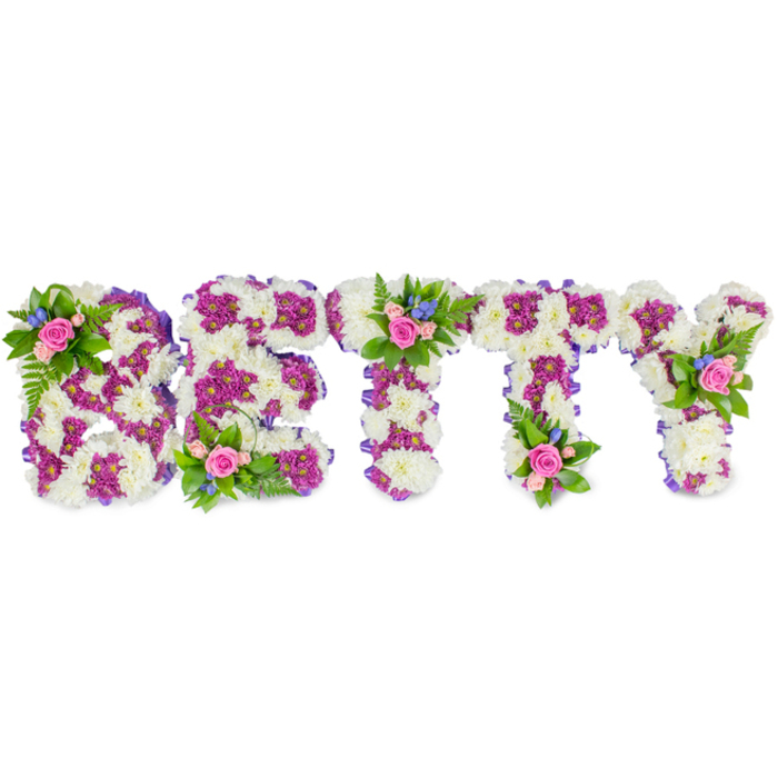 Presented horizontally against a spotless white field, this custom memorial piece spells the name BETTY in solid, capital floral letters that appear almost sculptural. Each letter's primary surface is a dense field of ivory-white chrysanthemums or similar creamy blooms, their clustered petals creating a velvety, tactile plane that invites a gentle touch in memory. Contrasting bands of lively purple and deep magenta flowers-small chrysanthemums or carnations-define inner forms and outlines, producing a graphic yet tender pattern that helps each character read clearly from a distance. The sides of each letter show a neat purple fabric or ribbon trim that softens the transition between face and edge and adds a poised, tailored look. Carefully positioned on top of the letters are three modest floral clusters: the upper left loop of the B, the crown of the first T, and the upper arm of the Y. These accent groups combine pale pink roses-some tightly budded, some open to reveal layered petals-with tiny azure-blue filler blooms (possibly forget-me-nots or small delphiniums) and sprigs of lush green foliage such as fern fronds, giving a sympathetic garden note. The roses introduce a delicate scent that complements the gentle, clean aroma of chrysanthemums while the blue flowers provide a soft visual breath. Photographed under even, diffused light, the arrangement projects quiet dignity and comfort appropriate for funeral services, wakes or memorials held by families in Queensbury and nearby Harrow, expressing personalised love and remembrance through thoughtful floristry.