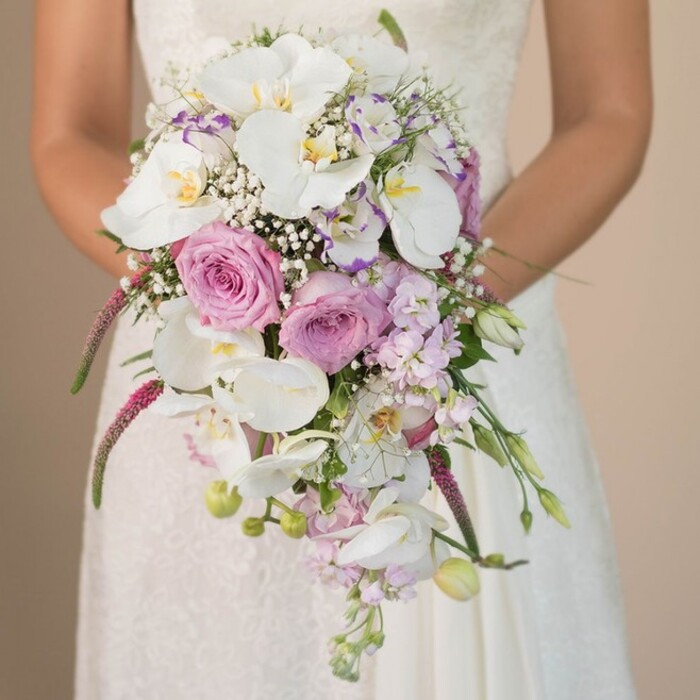 This bridal bouquet, captured in a peaceful Queensbury setting, reflects a floral masterpiece created with care and local flair. At its heart, velvety lavender roses unfold in quiet splendor, encircled by the serene elegance of white Phalaenopsis orchids whose broad petals and golden centers radiate luxury. Trails of magenta Amaranthus and wispy baby's breath interlace, blending structure with gentle whimsy as they spill in a graceful cascade. Interspersed with these are smaller lavender blossoms-perhaps stock or sweet pea-lending layers of colour and texture reminiscent of a stroll through Queensbury's local gardens in spring. Slender green tendrils and unopened buds peek through the blooms, adding natural freshness and depth. The bride's ivory gown hints at tradition with its lace overlay, complementing the bouquet's classic-meets-contemporary style. Subtle light falls on the bouquet, enhancing the interplay of white, pink, and purple-colours chosen for their serenity and romantic symbolism. The effect is both harmonious and dynamic, capturing the anticipation and joy that define a wedding day. As a local florist, seeing such a bouquet carried in weddings across Queensbury is a source of pride, as it brings together craftsmanship, beautiful materials, and the spirit of a community celebrating love, new beginnings, and the elegance of flowers.