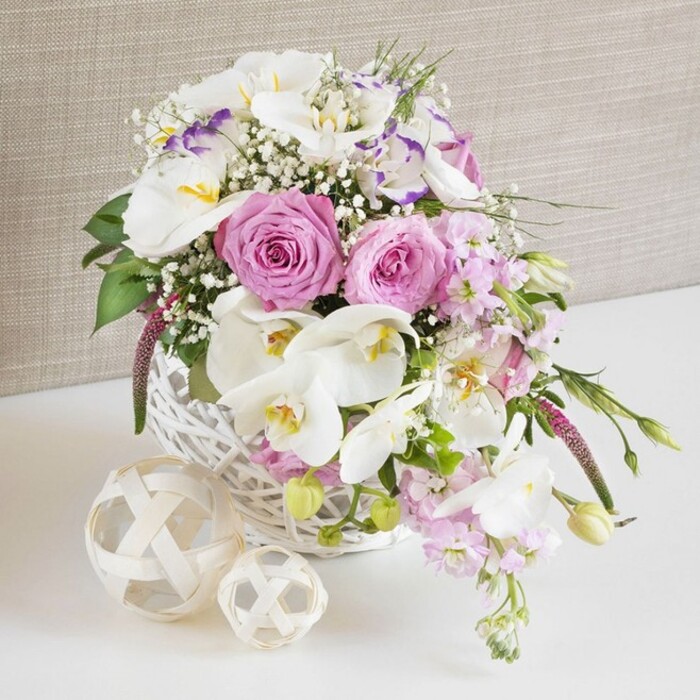 A beautifully styled floral composition fills the frame, evoking the feel of an intimate wedding table set up in a Queensbury townhouse or a small local restaurant. The focal point is a round white wicker basket, its tight weave and slightly matte finish giving a subtle, tactile charm. Overflowing from this basket is a lush mix of pastel pinks, creamy whites, and touches of soft purple. Several large pink roses, fully opened and perfectly formed, sit proudly at the center; their petal spirals are densely layered, creating a sense of depth and classic romance. Nestled around them are white Phalaenopsis orchids, each bloom broad and smooth, with glowing yellow-orange centers that draw the gaze inward and hint at an exotic, high-end feel often chosen for weddings. Interspersed are white Lisianthus flowers, some with delicate lavender edging that looks as if brushed on by hand, adding quiet sophistication. Tall stems of pale pink stock emerge and tumble slightly over the edge, suggesting gentle movement and a whisper of floral perfume in the air. Fine, cloudlike clusters of white baby's breath scatter between the larger blooms, lightening the overall look and giving a soft-focus effect. Sprigs of glossy green foliage and a few tender shoots trail from the sides of the basket, breaking the round silhouette and adding a natural, garden-fresh impression. On the bright white surface in front of the arrangement sit two decorative spheres crafted from narrow, intertwined strips of natural-colored material, echoing the woven basket and reinforcing the organic, artisanal theme. The background fades into a warm, neutral-toned fabric, slightly textured like linen or soft wallpaper, which keeps the viewer's attention firmly on the flowers. The entire scene feels poised for celebration, ideal as a wedding table centerpiece or engagement dinner arrangement for couples celebrating in and around Queensbury and nearby Kingsbury, with flowers that could easily have been hand-delivered that morning.
