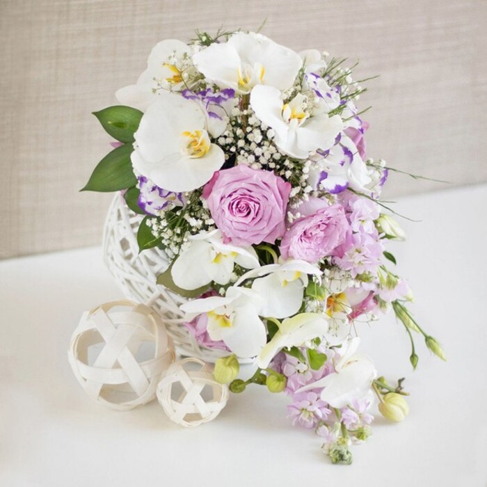 A carefully composed wedding-style table arrangement is centered on a white surface, its rounded silhouette framed by a gentle, light beige background that fades softly out of focus. The eye is first drawn to the sculptural vase, a perfect white sphere made from fine, interlaced strands that resemble hand-woven rattan or yarn, creating a modern yet delicate base. From this airy vessel rises a profusion of flowers in white, pink, and lavender tones, all arranged to create a sense of fullness and quiet luxury. Tall stems of white Phalaenopsis orchids arc elegantly, their broad, satin-like petals catching the light, each bloom showing a small splash of yellow and pale gold in the center that adds warmth and sophistication. Around these orchids, roses in muted blush pink and soft lilac are positioned at varying heights and stages of bloom; some are fully open, showcasing tightly layered hearts, while others are slightly cupped, promising further unfolding. Between them, smaller ruffled flowers, likely Lisianthus or Eustoma in white with purple-tinted edges, offer intricate detail and a gentle transition between tones. Airy clusters of white baby's breath are tucked throughout, giving the arrangement a light, clouded halo and preventing any single bloom from feeling too heavy. Slender, trailing stems of pale purple stock and similar blossoms spill naturally downward, echoing waterfall-style bridal work and guiding the eye over the edge of the vase. Deep green leaves and sprigs of foliage peek through to anchor the pastel palette. To the side, two cream-colored woven spheres of different sizes rest casually on the table, repeating the lattice pattern of the vase and suggesting considered styling for a reception space in Queensbury, perhaps in a local hall or restaurant near Kingsbury Road. The whole arrangement feels poised between contemporary design and classic romance, perfect for an elegant event or refined home décor.
