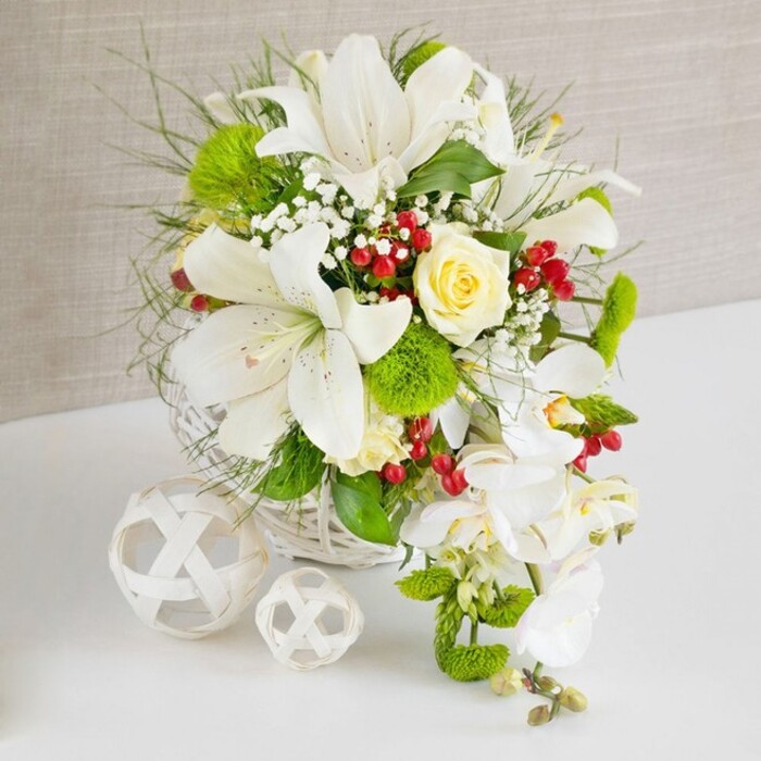 A sophisticated wedding-style arrangement rests on a clean white surface, carefully composed in a round, white woven basket that suggests both craftsmanship and comfort, much like the calm residential streets around Queensbury. The backdrop is a softly textured beige fabric, understated and warm, allowing the flowers to command all attention. Dominating the composition are several large white Oriental lilies, their petals fully open and curling slightly at the edges, revealing speckled throats and golden-tipped stamens that convey a sense of fragrant opulence. Near the top centre of the design, a single creamy ivory rose sits nestled among the blooms, its layered petals forming a gentle spiral that draws the eye inward. Around it, delicate sprays of white gypsophila float like mist, softening the outlines of the larger flowers. Vivid green pom-pom flowers, possibly Santini chrysanthemums or green dianthus, are dotted throughout, their rounded, tufted heads introducing a playful, tactile contrast against the sleek lily petals. Fine feathery foliage and slender blades of ornamental grass stretch outward and upward, adding motion and airiness, as if the bouquet were catching a slight breeze from Queensbury Park. From the right side of the basket, white phalaenopsis orchids spill in a gentle cascade, their arching stems and smooth, moth-like petals creating a waterfall effect that feels ideal for a wedding top table or engagement celebration. Interspersed among the greenery, clusters of rich red hypericum berries gleam like tiny jewels, adding depth and a subtle festive note. To the left, two white wicker spheres, one medium and one smaller, rest on the tabletop, echoing the basket's texture and balancing the arrangement visually, completing a scene of refined, celebratory elegance ready for local Queensbury delivery.