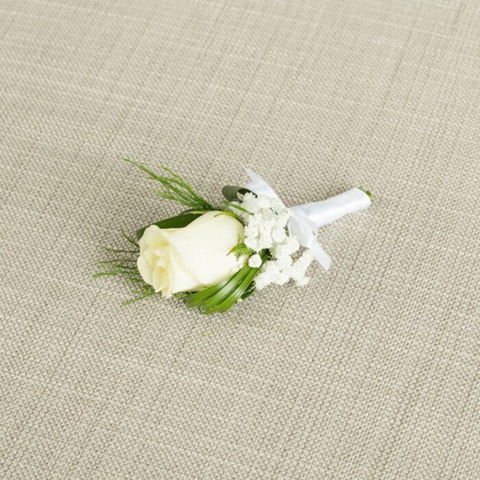 On a neutral, light beige background that resembles a cross-woven linen cloth, a single white rose boutonniere is displayed at a gentle angle, as though it has just been laid down by a florist making final checks before a Queensbury wedding. The rosebud at the centre is a soft, creamy white with a touch of ivory warmth, its petals tightly layered yet with just enough opening to show the classic spiral shape at its heart. At the base, faint green sepals peek out, hinting at the freshness of a bloom that has only recently been cut and prepared. Surrounding and slightly offset to one side are wisps of baby's breath, each stem branching into a constellation of tiny white blooms that create a delicate, cloud-like halo around the rose, giving the arrangement lightness and a sense of movement. Supporting the flowers are sleek, dark green leaves with a smooth, almost satiny finish and a few finer, fern-textured sprigs that lend detail and depth, much like the foliage often foraged or selected to echo the greenery found in Queensbury's small parks and garden borders. All stems are bound together with a crisp white satin ribbon, tightly wrapped in clean, even bands to hide the joins and give a comfortable surface for pinning to a jacket. Just below the rose, the ribbon gathers into a small, symmetrical bow, its loops and trailing ends lying flat and intentional. Soft, diffused lighting washes gently over the scene, revealing the subtle weave of the fabric beneath and the natural contours of each petal and leaf. The overall impression is of a timeless, elegant boutonniere designed for grooms, groomsmen, or formal guests at a refined event in or around Queensbury, capturing the quiet emotion and polished detail of a carefully planned wedding day.