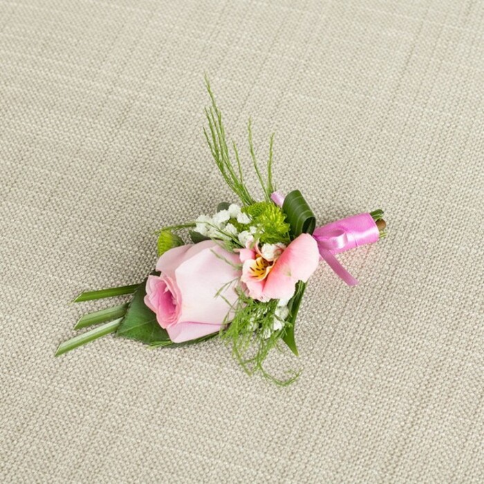 Gracefully styled and brimming with local character, this floral boutonniere lies diagonally across a richly textured beige fabric backdrop, perhaps evocative of a tablecloth found in a Queensbury family home. The composition is anchored by a tightly furled rosebud in the gentlest of pinks, its spiral pattern and tonal layers offering a portrait of delicacy and new beginnings. Nestled around the rose, an opening Alstroemeria blushes in soft peach, its petals radiating outwards to showcase miniature flecks of yellow and inky veins near the center-a signature flourish for thoughtful, bespoke designs from experienced Queensbury florists. Wispy sprigs of baby's breath impart a dreamy, cloud-soft levity, while small, round green cushion mums inject vibrancy, reminiscent of manicured garden hedges in the area. Framing the blossoms, knifelike blades of deep green foliage arch upwards, balanced by broader, structural leaves, some bent to cradle the flowers, others wrapped securely at the stem. The boutonniere is gathered and held fast by a splash of glossy fuchsia pink satin ribbon, tied off in a neat bow-its vivid colour promising festivity. The neutral, tactile fabric below enhances every shade and contour, inviting the hand to linger. Ideal as an accent for a wedding, prom, or treasured celebration in Queensbury, this compact design blends freshness, romance, and timeless style-perfect for lapels on momentous days or articles spotlighting local event floristry.