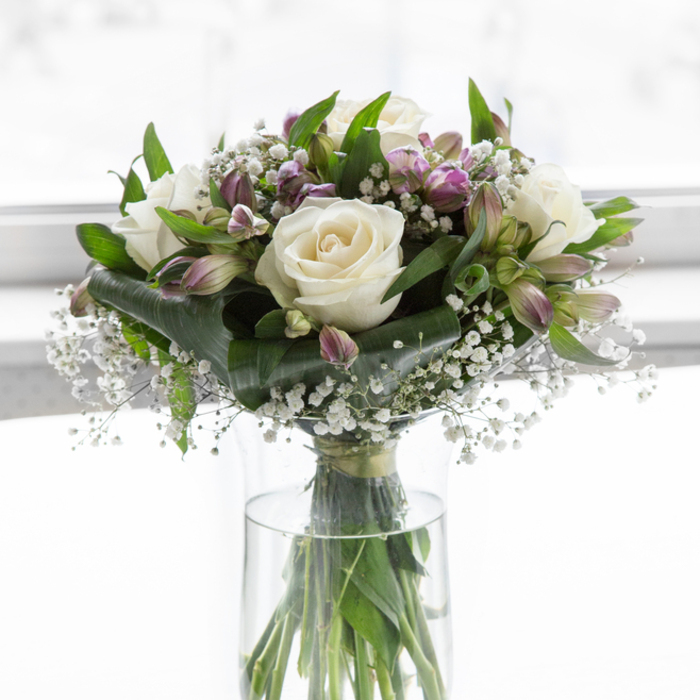 In this thoughtfully arranged sympathy bouquet, a cluster of creamy white roses takes centre stage, each bloom unfolding in soft, velvety layers that catch the light with a quiet, luminous glow. They are surrounded by graceful stems of alstroemeria, their petals painted in muted purples and lilac tones with faint touches of green at the edges, giving the arrangement a subtle wash of colour without disturbing its tranquil mood. Throughout the bouquet, fine sprigs of white baby's breath add a mist-like texture, filling the spaces between larger flowers so that no area feels empty, yet nothing appears crowded or overwhelming. The greenery is rich and plentiful, with glossy leaves and slender stalks framing the paler blooms and adding natural depth, much like the layered greens you might see in the gardens sprinkled around Queensbury. All of this rests in a clear glass cylindrical vase, where the straight, clean stems can be seen through crystal-clear water, underscoring a sense of freshness and care in the way the flowers have been prepared. The background appears very bright and softly blurred, suggesting a calm indoor setting in strong natural light-perhaps near a living room window on a quiet residential road off Salmon Street. This setting allows every petal, leaf, and stem to be fully appreciated, even in memory. The overall effect is one of serenity, tenderness, and respectful elegance, an arrangement that feels entirely appropriate for condolence, funerals, or placing in a peaceful corner as a lasting gesture of support and remembrance.