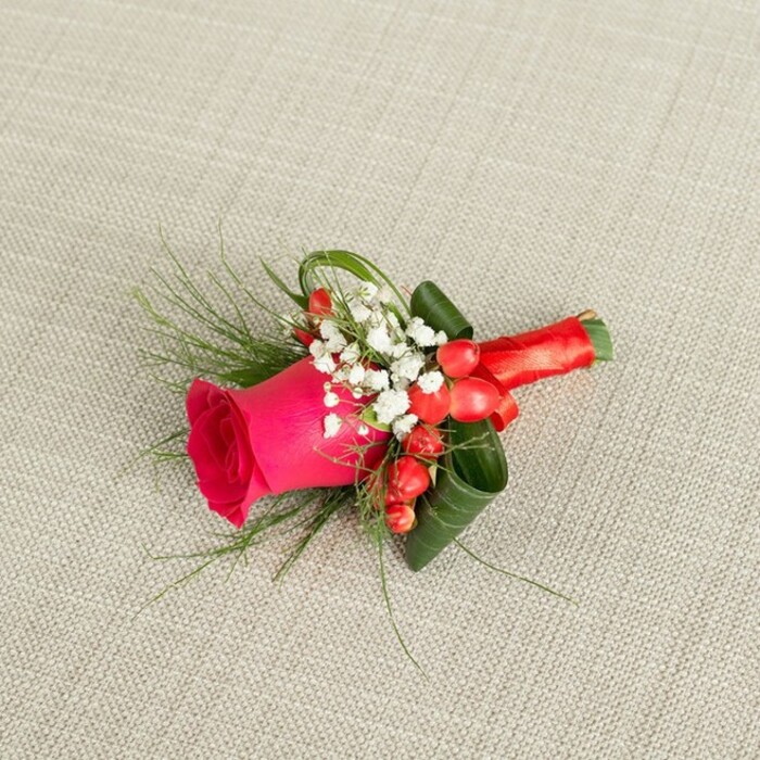 A small yet sophisticated floral boutonniere rests at a slight angle on a neutral light beige fabric backdrop, whose subtle woven texture suggests a refined linen, similar to the textiles often used in Queensbury's intimate wedding venues and community halls. The design is centred on a single striking fuchsia-pink rose, its petals packed densely in the middle and progressively loosening toward the edges, giving the bloom a lush, velvety appearance full of life and promise. Encircling this rose are ethereal clusters of white baby's breath, the tiny, cloud-like blossoms creating a soft halo that brings gentle lightness and a classic bridal feel to the piece. Adding contrast and festive energy, several bright red hypericum berries are nestled close to the rose, their smooth, rounded surfaces catching the light like little jewels. Surrounding and supporting the flowers is an artful mix of greenery: fine, wispy fronds, likely asparagus fern, stretch out delicately, while wider, deep green leaves are carefully folded and curled, forming an architectural cradle that frames the central rose. The stems are tightly bound with a rich red satin ribbon, wrapped with precision and tied in a small, tidy knot that keeps the profile slim and comfortable for wearing on a lapel. Soft, even lighting falls across the arrangement, highlighting every texture-from the matte petals and airy white blossoms to the sleek shine of berries and ribbon. The overall impression is of a beautifully balanced accessory, crafted by an experienced florist for a groom, best man, or prom guest in Queensbury, capturing romance, joy, and understated elegance in a single, handcrafted detail.