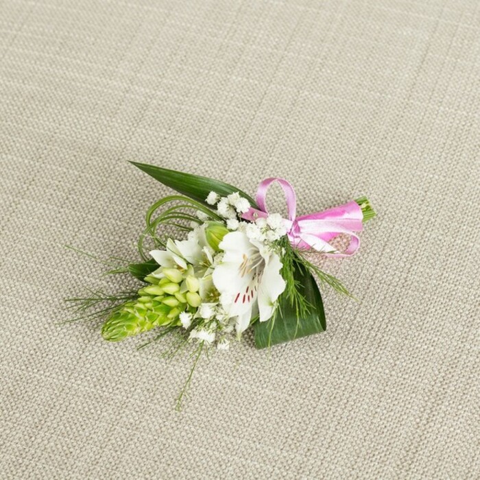 A refined, compact boutonniere is displayed against a softly textured, light beige fabric background, similar to natural linen, whose understated tone allows the floral details to shine. At the centre of the composition is a single white alstroemeria, sometimes called a Peruvian lily, its petals opening generously to show off a pattern of fine reddish-brown speckles and streaks concentrated near the throat. The petals appear smooth yet gently rippled at the edges, with their creamy whiteness subtly warmed by the even light that bathes the arrangement. Nestled beside the alstroemeria is an eye-catching spike of bright lime-green buds, reminiscent of a compact Ornithogalum cluster, each tiny bud tightly folded to create a sculptural, conical form. Threaded throughout are sprigs of delicate white baby's breath, their minute, cloud-like flowers softening every transition and lending the piece an ethereal, almost floating quality. The blooms are supported by a mix of lush greenery: broad, dark green leaves provide structure and depth, while finer, grass-like blades arc outward, introducing movement and a slightly whimsical feel, as if caught by a gentle Queensbury breeze along a quiet residential street. On the right side, the stems are gathered neatly and finished with a small bow of glossy pink satin ribbon, its bright, joyful colour adding a subtle note of festivity and romance. The lighting is even and flattering, revealing the textures of petal, leaf, and ribbon with clarity. This boutonniere feels perfectly suited to a Queensbury wedding, registry ceremony, or prom, a carefully crafted flourish that would sit gracefully on a lapel or wrist, reflecting the thoughtful craftsmanship of an experienced local florist.