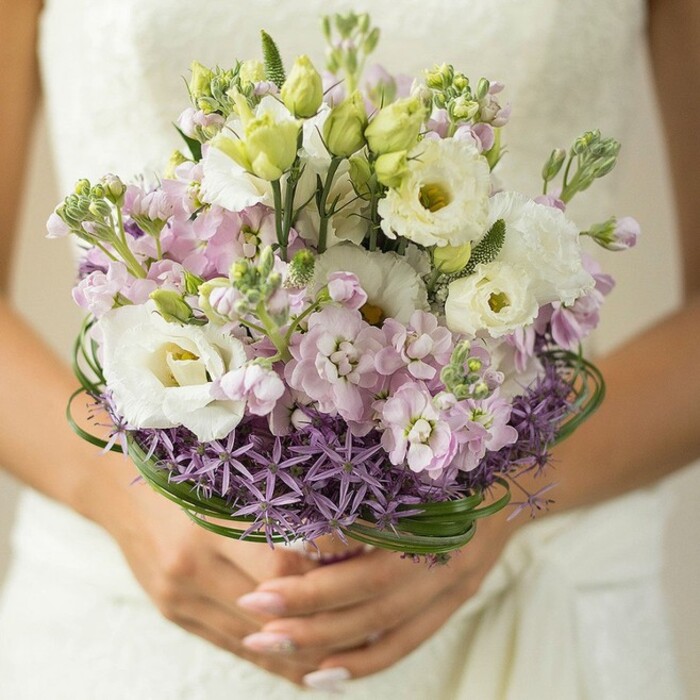 Captured in exquisite close-up near Queensbury's leafy boulevards, this image centers on a lush, artisanal wedding bouquet, carefully arranged and held by a bride whose hands support the composition with a delicate grace. Her nails, finished in a natural blush or soft pink, further accentuate the bouquet's romantic palette, while a glimpse of an ivory lace dress forms a gentle, blurred backdrop-an allusion to time-honored wedding traditions in north-west London. The arrangement itself unfolds in romantic layers: majestic white lisianthus with ruffled petals and subtle green-yellow hearts are interwoven with petite clusters of fragrant lilac stock blossoms, gently inviting both eye and nose to linger. Slender unopened blooms and glossy greenery offer refreshing accents and visual depth, while the foundation of spiky, star-form purple allium at the bouquet's base draws the gaze with its modern architectural flair. Encircling the whole are refined loops of deep green bear grass, their curving lines adding sophisticated movement to the silhouette. Subtle light plays across each petal, highlighting the blend of pastels and creating an effect both airy and grounded-the very spirit of a Queensbury wedding morning. This bouquet, prepared by expert local florists, speaks of gentle romance, vibrant freshness, and the timeless artistry of hand-tied wedding flowers, perfect for stories that celebrate love, community, and the tranquil beauty of a special day spent in Queensbury.
