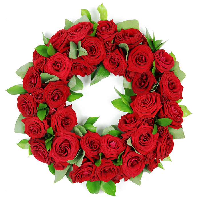A deeply symbolic circular wreath of red roses rests against a pristine white background. The wreath is densely packed with dozens of crimson blooms opening at various stages, from tightly furled buds to elegantly unfurled velvety petals, each displaying rich hues and intricate folds. The abundant red roses, traditionally symbolizing love, remembrance, and respect, create a continuous unbroken ring. Interspersed among the blooms are clusters of fresh glossy green foliage. Some leaves are broad and rounded, possibly eucalyptus, while others are slender and pointed, offering verdant contrast that enhances depth and natural beauty. The careful arrangement gives the wreath a full three dimensional quality, with petals and leaves subtly overlapping. This elegant red rose wreath conveys a message of solemn beauty, heartfelt emotion, and enduring tribute. It is a powerful visual element often associated with memorial services, funeral arrangements, expressing deep sympathy and condolences, or honoring anniversaries and commemorations. The striking combination of passionate red and serene green set against a pure white backdrop makes this wreath a poignant representation of remembrance and respect. The narrative feel is one of quiet dignity and artistry, suitable for publishing alongside articles about grief, memorials, or flower based tributes for the Queensbury community. It is available for delivery to local venues and homes in the area, with options for same day service across Queensbury.
