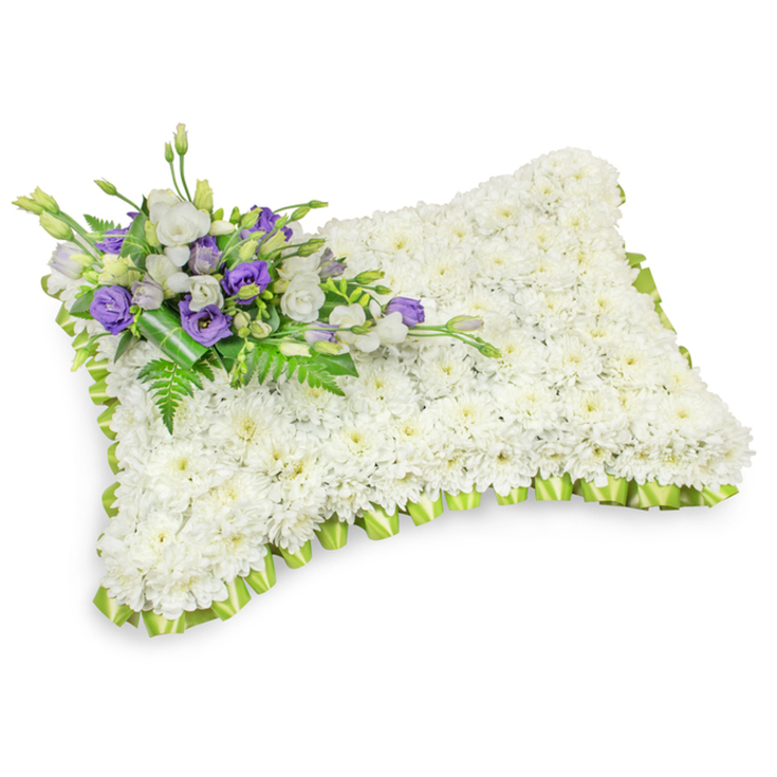 An exquisitely formed rectangular memorial pillow, composed almost entirely of compact, snowy-white chrysanthemum heads arranged so densely they create an even, cushion-like plane of floral purity. The overall silhouette is softly rounded at the corners with a subtle raised profile, visually and texturally reminiscent of a sleeping pillow meant to symbolize rest. The perimeter is framed by an artful light-green ribbon, pleated into a delicate scalloped border that provides a tender contrast and tidy definition against the immaculate white surface. At the pillow's upper-left corner an intentional spray of accent blooms interrupts the white expanse: layered white lisianthus, and royal purple lisianthus in various stages of bloom, bringing muted colour and a sense of solemn dignity; the purple tones traditionally suggest respect and reverence. Among these blooms slender fern fronds and glossy foliage are woven through, offering feathery texture and a living green counterpoint, while a wider dark-green leaf wrap secures the stems and lends a refined, handcrafted finish. Soft, neutral studio lighting and a subtle drop shadow emphasize the arrangement's three-dimensional form without distracting from its somber purpose. Presented with the quiet care of an experienced neighbourhood florist, this pillow reads as an appropriate tribute for funerals, wakes, or graveside services in Queensbury, near local landmarks such as Queensbury station and the green spaces around Fryent Country Park, gently conveying sympathy, remembrance and the careful touch of local floral tradition.
