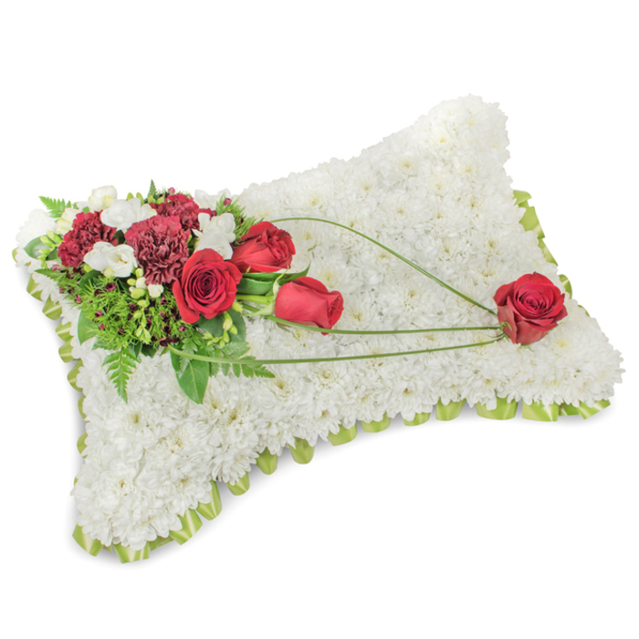 An elegant rectangular floral pillow rests composed on a clean white plane, its entire body formed from hundreds of small, immaculate white chrysanthemum blooms packed tightly together to evoke a plush, cloud-like surface. Along the slightly raised perimeter a pale green ribbon or delicate foliage edge registers as a soft outline, introducing a fresh, spring-like note against the snow-white field. On the upper left a lush cluster of deep red roses and dark burgundy carnations creates a rich, dramatic focal point; the roses show stages of bloom from compact buds to fully unfurled petals, while the ruffled carnations add texture and depth. Nestled among the reds are small white sprays - likely freesias or tiny chrysanthemum florets - that offer bright punctuation and a subtle, clean fragrance. Greenery provides the supporting architecture: slender fern fronds, salal-like leaves and long, glossy stems give density and contrast. From the main spray three elegant, sweeping lines of light green foliage or bear-grass extend diagonally across the white chrysanthemum surface toward the lower right corner, guiding the eye with graceful arcs. A single, perfectly formed red rose rests where those lines terminate, appearing almost cradled by the bend of the foliage and symbolising lasting love and remembrance. The composition reads as a calm and respectful sympathy cushion, appropriate for funeral services and memorial displays in Queensbury and the surrounding communities near Queensbury tube, Fryent Country Park and the high street. The combined textures, subtle scent and considered movement of the design make it a timeless tribute of comfort and honour.