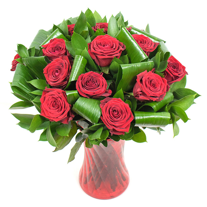A classic yet contemporary bouquet of deep crimson roses is shown here in full, sumptuous bloom - more than a dozen heads forming a generous, rounded silhouette that reads as both abundant and carefully restrained. Each rose displays densely spiralled, velvety petals that darken toward the center and soften toward their outer edges, catching the studio light in subtle satiny highlights. Interspersing the blooms is a lavish quantity of dark green foliage: long, glossy leaves create a thick, verdant foundation while broader, emerald-hued leaves have been artfully curled and spiralled into elegant loops, their sculpted forms offering striking contrast to the natural softness of the roses. The stems and greens are held inside a tall, translucent ruby-red glass vase with a delicate ribbed or twisted surface; its vertical texture reflects light in faint bands and echoes the bouquet's verticality, while the vase's rich colour complements the roses without overwhelming them. Photographed against a pristine white background, the composition isolates the flowers' intense reds and deep greens, making the tactile differences - velvet petals, glossy foliage, smooth glass - immediately apparent. Picture this arrangement arriving at a family anniversary gathering in a house tucked off Queensbury's main roads, or being handed over after a thoughtful order placed by a neighbour on Kingsbury Road; the piece carries the quiet confidence of experienced local floristry. The overall mood is one of enduring passion and refined taste, a floral statement suitable for romantic milestones, special celebrations, or elegant home décor.