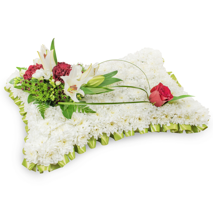 Against a spare white background, this elegant pillow-shaped sympathy arrangement is composed of a compact sea of tiny white chrysanthemums, each bloom nestling so close to its neighbours that the surface appears like a plush, serene textile. The effect is tactile and calming: the chrysanthemums' fine petals suggest softness and repose. A neatly pleated ribbon in a pale lime-green tone outlines the rectangle, its soft sheen and slight scallop lending refinement and a whisper of colour along the edge. At the upper left, a natural cluster forms the emotional heart of the piece - two alabaster Oriental lilies fully open to reveal their warm yellow stamens, an awaiting lily bud, and clusters of deep burgundy spray carnations that provide a dramatic, respectful contrast. Varied greenery, including feathery fern fronds and broader leaves, supports and frames these focal blooms, adding depth and a sense of living texture. From this grouping, a slim arch of green salal or decorative wire travels gracefully to the right, terminating in a single deep red rosebud poised on the pillow's upper right - its velvety petals just beginning to part, signalling enduring love and quiet remembrance. The carefully balanced palette of white, soft green and rich red, paired with gentle studio light, conveys dignity and tranquillity. As a local florist serving the Queensbury area and surrounding neighbourhoods, I would recommend this arrangement for funerals, memorials, or bedside tributes where comfort and respectful presence are needed.