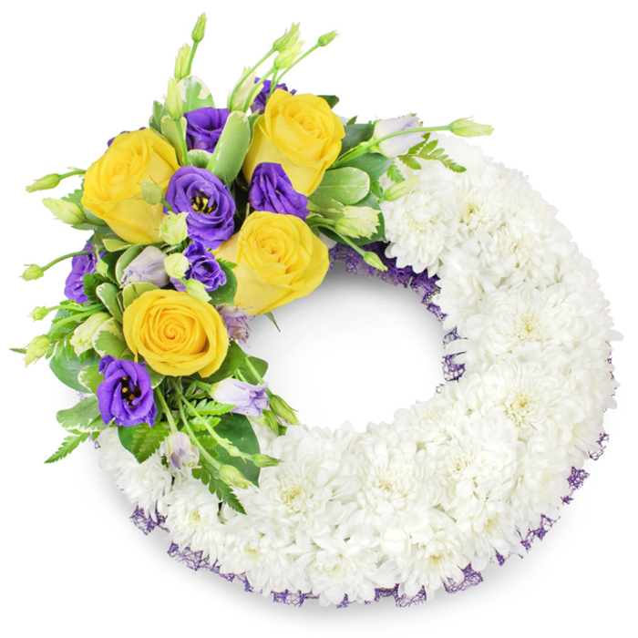 Presented against a clean white surface, this circular sympathy wreath reads as an expert study in contrast and comfort: the main body is composed almost entirely of tiny white spray chrysanthemums, packed closely to form a continuous, plush ring whose many layered petals create a soft, almost cushion-like sweep. The whites are cool and pure under the light, the surface appearing almost clouded and whisper-quiet. On the upper left hand a composed floral spray introduces warmth and depth - several honey-yellow roses with petals gently unfurling, radiating a subtle, honeyed scent memory, sit beside clusters of deep purple lisianthus whose velveteen, ruffled blossoms add a stately, contemplative mood. A smattering of pale lavender and green Eustoma buds punctuates the cluster, providing visual cadence and the gentle optimism of things yet to bloom. Foliage choices - glossy, rounded leaves, some with faint variegation, and fine fern fronds - weave through the arrangement, offering lively green notes and a stabilising structure. A slender ribbon or mesh in dark purple peeps from beneath the mass of white along both the inner and outer edges of the wreath, a discreet detail that echoes the lisianthus tones and refines the silhouette. This is a design often requested for funerals at local chapels and community memorials around Queensbury and the Jubilee line corridor; its balance of purity and colour communicates condolence, dignity, and a careful, handcrafted attention to composition.