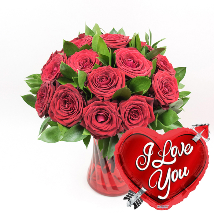 An opulent bouquet of deep red roses is showcased against a spotless white background, the contrast drawing the eye immediately to the rich colour and texture of each bloom. Around a dozen or more roses are nestled tightly together, creating a dome of petals that looks both luxurious and inviting. The flowers range from almost fully open blossoms, their velvety petals curling softly outward, to more modest buds still in the process of unfolding, lending the arrangement a sense of natural rhythm and progression. Between the blooms, fresh green leaves weave through the composition, their glossy surfaces catching the light and framing the roses with a lively, verdant edge. The bouquet rests in a clear, ruby-tinted glass vase with elegant, vertical fluting. These ridges in the glass subtly bend the light, sending streaks of red along the stems and into the water, and adding depth and texture to the otherwise smooth surface. The rich ruby tone of the vase feels particularly fitting for romantic deliveries across Queensbury, where a single, dramatic piece like this can brighten a home on a quiet evening. To the lower right of the arrangement, slightly overlapping the vase, a bright red heart-shaped foil balloon adds a playful finishing touch. Its shiny metallic sheen reflects the whites and reds around it, and in the centre, the phrase I Love You appears in crisp, white cursive script. A slender silver Cupid's arrow pierces the heart diagonally, transforming the balloon into a familiar emblem of romantic devotion. Together, the roses, foliage, vase, and balloon form a classic love token, ideal for anniversaries, Valentine's Day, or anytime a grand gesture of affection is needed.