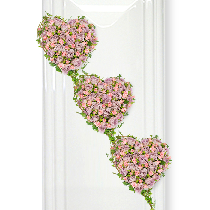 This image shows a beautifully detailed funeral tribute composed of three interconnected hearts made entirely of miniature roses, arranged so they appear to cascade gracefully from the top left to the bottom right of a clean, white background. Each heart form is lush and full, its surface covered in small to medium rose blooms in a harmonious mix of dusty lavender, soft mauve, and gentle pastel pink. The roses are placed tightly together, creating a dense, velvety carpet of petals that looks both luxurious and tender, with some blooms fully open and others just beginning to unfurl, suggesting the passing of time and the stages of a life well lived. Defining the outer edge of each heart, and flowing naturally between them, is a line of fresh green ivy. The ivy's slender stems and polished, deep-green leaves encircle the roses and then trail downward, linking each heart to the next so the entire design feels like one continuous, living garland. The contrast between the cool greens and the soft rose tones gives the arrangement a calm, soothing rhythm, like a quiet walk through a leafy corner of Queensbury on a still morning. Soft, even lighting ensures every petal and leaf is visible without harsh shadows, while faint vertical reflections on the left side hint at a glass or glossy surface, subtly suggesting a tasteful, modern interior such as a chapel or funeral home reception near Queensbury underground station. This cascading trio of hearts is clearly designed as a heartfelt tribute, ideal for expressing deep affection, gratitude, and remembrance during a funeral or memorial gathering in the Queensbury community.