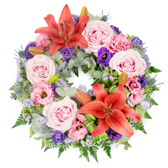 Viewed from directly above against a crisp white backdrop, this funeral wreath forms a perfectly even circle, dense with flowers and foliage that evoke both comfort and quiet strength. Two standout coral-tinged, reddish-orange Asiatic lilies serve as the main points of focus, one gracing the upper arc of the wreath and the second situated just off-centre towards the bottom, their broad petals fully unfurled and stamens clearly visible. Between and around these lilies, numerous blush to lavender-pink roses create a soft, romantic layer, their gently spiralling petals suggesting tenderness and enduring affection. Interlaced among the roses are lisianthus blooms in shades of delicate pink and rich purple, their ruffled, cup-shaped flowers adding movement and subtle shadows that deepen the composition. Sprays of tiny lavender-blue filler flowers, likely statice, nestle between the larger heads, giving the impression of a light, airy haze. Spiky blue eryngium thistles punctuate the greenery at intervals, their cool blue-grey tones and bristled texture lending an almost sculptural quality that contrasts beautifully with the softness of the roses and lisianthus. The foliage base is abundant and varied, including rounded, silvery eucalyptus leaves, slender fern fronds, and darker, glossy greenery that fills the circle without any gaps. Several closed lily buds lie quietly among the leaves, symbolising life continuing beyond the moment of farewell. Near the lower lily, folded and looped bright green leaves, likely aspidistra or Ti, introduce a touch of modern design, highlighting the care and precision typical of experienced florists in Queensbury. This wreath would be a thoughtful choice for a memorial or funeral service, its harmonious palette and balanced form expressing respect, remembrance, and a gentle sense of peace.