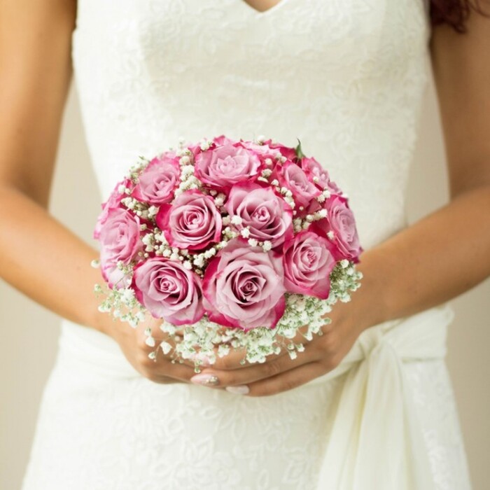 The heart of a Queensbury wedding is beautifully distilled in this intimate, detailed image-a bride caught in a moment of stillness, hands gracefully resting just below her waistline, drawing the eye towards the centerpiece: her exquisite bouquet. Here, the florists' careful artistry shines in every detail. A perfectly rounded dome of roses bursts with lush colour, each petal celebrating the full spectrum from barely-there blush at the center to vivid, fuchsia-tinged edges. The dual-tone blooms create a lush sense of depth, reminiscent of the garden blooms found along Church Lane. Scattered between them, dainty sprays of baby's breath bring a frothy, cloudlike softness, lending a timeless romantic feel much loved in suburban weddings. The bouquet is held with care in hands featuring a gentle, sun-kissed tone and delicate, pearly manicured nails, echoing the classic style of many brides passing through Queensbury's charming chapels. The sleeveless dress, with its floral lace bodice and understated sash, feels both traditional and unmistakably personal. Soft, diffuse natural light enhances the white, beige, and pinks, bringing life and warmth to the composition. For those planning a wedding in Queensbury or searching for the perfect floral touch, this image perfectly encapsulates the emotional significance and meticulous detailing that define a truly memorable bridal bouquet.