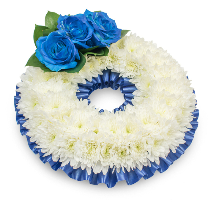 This elegant wreath, photographed from a slightly raised angle against a spotless white field, combines traditional symbolism with composed modernity to form a respectful memorial piece. The main body is a continuous band of immaculate white chrysanthemums, densely arranged to create a soft, slightly domed surface whose countless feathery petals give the impression of a gentle, breathable cloud. Tiny hints of creamy yellow at the flower centres temper the coolness of the white, adding a nuanced warmth. Anchoring the upper left quadrant are three vivid, deep-blue roses whose velvety, tightly coiled petals present an intense, jewel-like colour against the white. The roses are set amid a small spray of dark green foliage that provides both contrast and a natural silhouette. A pleated satin ribbon - the same rich cobalt hue as the roses - outlines the wreath on both inner and outer rims; its ruffled texture and luminous sheen frame the flowers and lend a finished, dignified edge. The wreath casts a faint shadow on the background, which gives depth and a subtle feeling of presence, suitable for display at a chapel, funeral parlour, or graveside. From a florist's perspective, the scent is quietly reassuring - the lightly herbal note of the chrysanthemums supporting the soft perfume of bloom-sweet roses. Locally, families in Queensbury and neighbouring Kingsbury or Wembley often choose such a combination for its respectful symbolism and understated beauty, using it as a lasting token of remembrance at memorial gatherings and services.