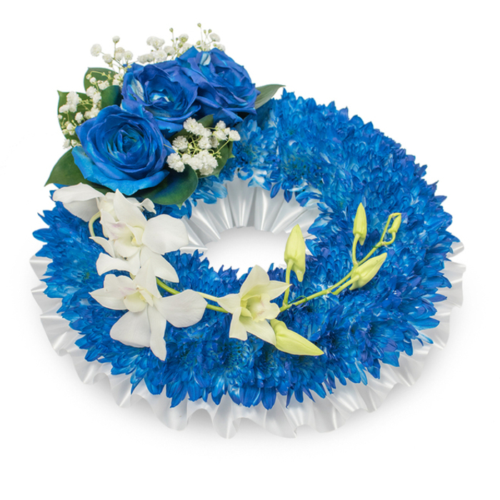 A dignified memorial wreath, rich in blue and white, presented on a clean white field so that every floral detail is visible: a full ring of small, royal-blue chrysanthemum-like flowers forms the bulk of the piece, their tightly packed petals creating a deep, textured surface like a sea of indigo. The ring's inner edge is lined with a pleated white satin ribbon, its subtle sheen and ruffled contour framing the vibrant blue and giving the wreath a classic, polished appearance. Anchoring the design at the upper-left curve is a small cluster of three large blue roses, petals unfurled in a deep sapphire hue with a velvety finish; they are accented with tiny white baby's breath and touches of dark green foliage, lending a timeless, respectful air. A slender spray of pristine white orchids - resembling Phalaenopsis or Dendrobium - cascades from mid-left down toward the lower-right, their sculpted petals and pale throats introducing a soft, floating contrast to the saturated blue. Several fresh green orchid buds along the stem suggest renewal amid remembrance. The arrangement is photographed under cool, soft light that highlights contrasts between the matte chrysanthemums, glossy rose petals, and the silk-like orchids. Crafted with care for memorial services and designed to sit gracefully at a chapel or grave-side near Kingsbury, Wembley or Harrow, the wreath communicates serene sympathy, enduring memory and the quiet beauty of a heartfelt farewell.