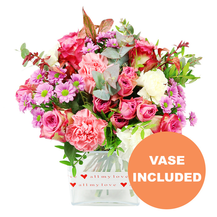 This image showcases a lush, carefully composed mixed bouquet that looks like it has just been finished on the workbench of a local Queensbury florist, then placed against a pure white background so every detail is clearly visible. Dominating the arrangement are multiple pink roses, some fully open to reveal layered, velvety petals in deep fuchsia, others still slightly cupped in softer blush tones. Intertwined with them are generous heads of carnations, their ruffled petals in coral, peach-pink and creamy white creating a plush, almost cushion-like texture. Sprinkled throughout are small daisy-style chrysanthemums in lavender and purple shades, each with a bright, lime-green centre that adds a spark of freshness and contrast. The flowers are nestled into a bed of mixed foliage: rounded eucalyptus leaves with their cool, silvery-green wash; darker green leaves that lend structure and fullness; and a few reddish-brown sprigs, which give a gentle nod to autumnal warmth without overpowering the romantic mood. Everything is gathered neatly into a rectangular clear glass vase with slightly rounded edges, the stems clearly visible through the water. Around the base of the vase is a white decorative band printed with tiny red hearts and the flowing phrase all my love, repeated like a soft refrain, making the bouquet an obvious choice for heartfelt occasions. In the lower right corner of the photo, an orange circular overlay boldly states VASE INCLUDED in white capital letters, highlighting that the design arrives as pictured, ready to place on a dining table in a Queensbury home, a desk in a local office on Mollison Way, or a bedside table for a special celebration.