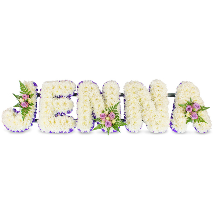 A lovingly assembled funeral tribute spells JENNA in clean, three-dimensional capitals formed from clusters of creamy white chrysanthemums, their compact petals creating a plush, cloud-like texture that feels both soothing and solemn. Each letter is crisply edged with a narrow band of rich purple ribbon or tiny lavender blooms, lending contrast and a quiet elegance to the otherwise immaculate white composition. Three artful accent posies punctuate the name: one placed within the upper left curve of the J, a second centred at the base of the second N, and a third sitting on the upper right curve of the A; these consist of soft pink and lavender spray roses, a few pale cream-yellow roses, and slender dark-green fern fronds that add depth and a delicate, ferny scent. Thin dark connecting rods subtly link the letters, lifting and stabilising the piece so it reads as a cohesive tribute on a memorial table or against a chapel wall. The photograph captures soft, even light falling across the florals, giving attention to petal detail, ribbon texture, and the gentle shadows beneath each letter. Flower Delivery Queensbury prepares tributes like this for funerals and memorials across Queensbury, Kingsbury and Wembley, where families seek a bespoke, respectful way to mark a life with a personalised floral name display that conveys love, reverence and lasting memory.