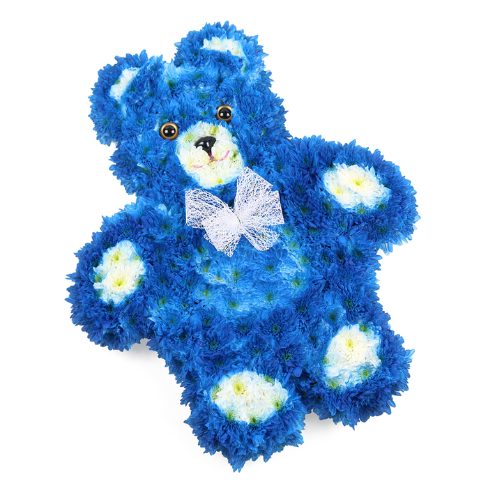 Set against a clean, uncluttered white background, a floral teddy bear tribute captures the eye with its intense royal blue colouring and tender detailing. The bear is designed to lie flat, its rounded silhouette and gently splayed limbs giving it the familiar shape of a cherished children's toy. Each part of its body is covered in tightly packed, small-petalled blossoms, likely chrysanthemum or similar blooms, creating a dense, cushion-like surface that mimics the softness of fur. The blue petals form the main structure - head, ears, arms, belly, and legs - while clusters of creamy white and soft yellow-tinted flowers highlight the inner ears, the pronounced snout, and the oval pads on each paw. These lighter areas bring a gentle radiance that softens the rich blue, catching the light and drawing focus to the bear's friendly features. The eyes are dark, round, and glossy, set close together for a childlike innocence, and the black triangular nose with a fine curved line beneath suggests the simplest of smiles. Around the neck, a fine white mesh or organza bow lies neatly, its delicate folds and transparent sheen adding an air of purity and ceremony. Imagined being prepared in a quiet Queensbury workshop, not far from the station or local churches, this floral teddy feels ideal as a sympathy tribute for a little one or as a comforting presence at a funeral service. The colours whisper of peace, serenity, and remembrance, offering a visual embrace to families in Queensbury who wish to send a message of love that is soft, respectful, and beautifully crafted.