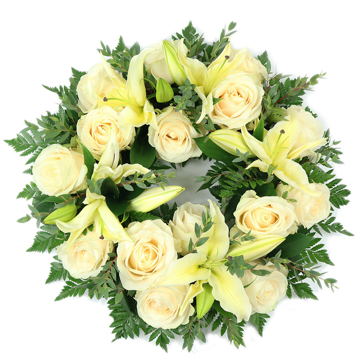 Viewed from above against a plain white plane, this elegant funeral wreath is a study in restraint and gentle comfort. The circular design is densely built, with some fifteen to twenty creamy ivory roses forming the principal rhythm of the piece; each rose shows the layered, spiralling petals that give the bloom a calm, ordered beauty. Scattered among the roses are soft, pale yellow lilies - a mix of fully opened trumpet-shaped flowers and several buds that lend a poised vertical accent - their pale stamens and graceful petal curves adding sculptural interest. The floral elements are set against a rich tableau of greenery: fine, feathery fern fronds trace the wreath's outer edge, creating a soft, natural halo, while darker, broader-leafed sprigs, likely eucalyptus, introduce cooler green tones and a modest textural contrast. The overall symmetry and fullness read as carefully measured and respectful; the wreath's unbroken ring symbolizes continuity, mourning, and hope. The lighting is neutral and soft, allowing the creamy tones to remain unvarnished and true, and giving the arrangement a dignified stillness. Sensory impressions imagine the roses' powdery sweetness tempered by a green citrus whisper from the lilies and the faint, aromatic freshness of eucalyptus. Designed for memorial services, graveside tributes, or as a tender expression of condolence, this wreath carries a composed, consoling message - an understated, living emblem of remembrance for families in Queensbury and surrounding communities seeking a graceful farewell.