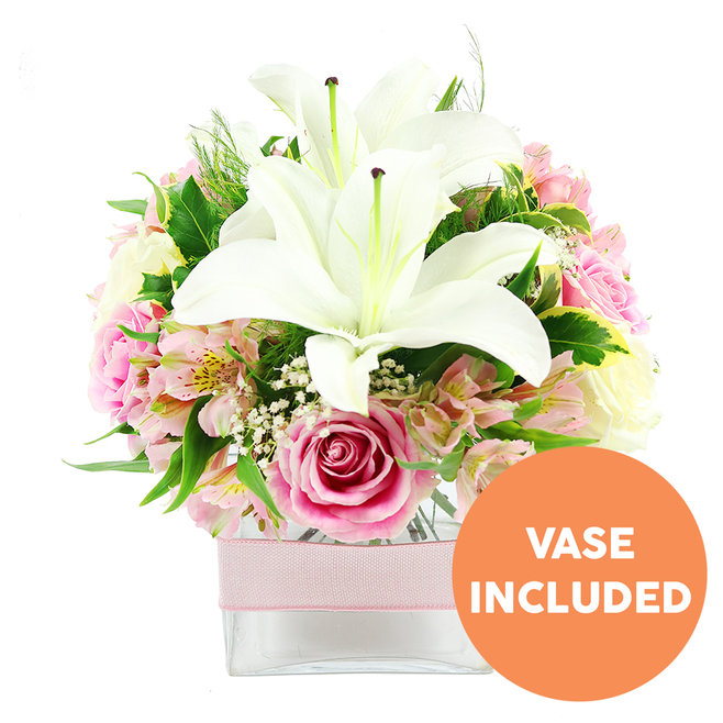 A beautifully balanced floral arrangement of white lilies and pink roses is shown in a low, square glass vase, photographed against a pure white backdrop that heightens the colours and textures. At the heart of the design, two fully open white Oriental lilies take pride of place, their large, trumpet-like blooms flaring outward with a sculptural grace. The lily petals are creamy white and slightly ruffled at the edges, and at their centers stand bold reddish-brown stamens, dusted with pollen, giving the impression of rich detail even at a distance. Encircling these lilies is a generous band of soft pink roses in many stages of opening, from tightly furled buds to wide, layered blooms that display each petal like the pages of a book. Among them, dainty stems of pink and white alstroemeria introduce fine speckles of yellow and subtle striping, adding a sense of depth and movement, while clusters of tiny white baby's breath flowers float lightly between the larger heads, creating an almost cloud-like softness. Varied greenery, including smooth, glossy leaves and finer, feathered foliage, forms a lush green cushion that supports and frames the flowers, reminiscent of the quietly tended shrubs and hedges lining residential streets around Queensbury Station. The arrangement sits firmly in a clear glass cube vase, the straight lines of the container contrasting with the soft curves of the petals. A pale pink ribbon tied horizontally around the vase adds a gentle, romantic detail. In the lower right corner of the image, an orange circular badge with crisp white lettering announces VASE INCLUDED, clearly explaining that the vase is part of the floral gift. The composition exudes calm elegance, perfectly suited to a coffee table in a Queensbury living room or as a welcoming centrepiece delivered to a local office on a special occasion.
