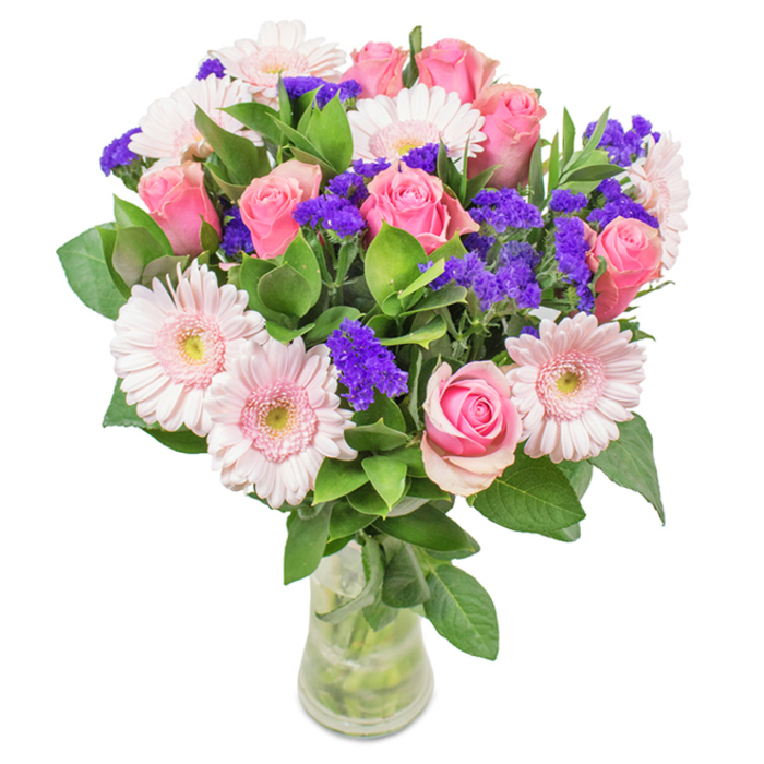 An exquisitely arranged bouquet, freshly delivered in Queensbury, stands poised in a crystal-clear glass vase against a pristine background, its colours radiating soft elegance. Central to this composition are approximately seven to nine dusky pink roses, the outer petals still gently furled or beginning to spiral open-each bloom meticulously selected for its velvety texture and timeless appeal. Interwoven amidst them, around five to seven pale pink Gerbera daisies broadcast their charm, their generous round heads and creamy, almost white centres creating a gentle luminosity that draws the eye. Interrupting this pastel harmony are vivid clusters of purple statice or limonium, whose unique, papery texture infuses the bouquet with both contrast and subtle drama. Lush, deep green foliage fans out around the base, lending abundance and a sense of the fresh gardens found tucked behind Queensbury's family homes. The slender glass vase reveals the cut stems and glistening water, reinforcing the bouquet's freshness-an ideal centerpiece for a birthday, anniversary, or heartfelt moment shared across Kingsbury or Edgware. Every aspect of this arrangement is crafted with sophistication in mind, celebrating joy, beauty, and the warmth of personal connection in Queensbury.