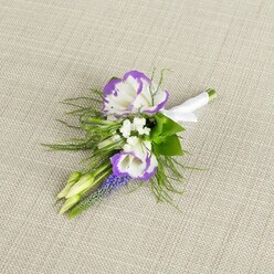 A single, thoughtfully composed boutonniere rests on a pale beige, finely woven fabric that resembles natural linen, the subtle texture beneath it providing a calm, almost tactile backdrop. The focal point is a pair of open lisianthus flowers, their white petals elegantly ruffled and edged with a vivid band of purple, like a soft watercolour wash that has settled along each curl of the bloom. Around them, small white filler flowers cluster in loose sprays, adding a misty lightness and an impression of tiny stars scattered among the petals. A slim spire of muscari rises beside the central blossoms, each tiny, bell-shaped floret a deep grape-purple, giving the arrangement a grounded vertical line and a slightly woodland feel, as if gathered from a garden not far from a quiet Queensbury side street. Wisps of fine, feathery greenery extend outward, their light, airy fronds softening the edges and evoking the gentle, green freshness that lingers after rain over local greens and footpaths. Two unopened buds, pale green and smooth, nestle discreetly amongst the foliage, suggesting new beginnings and the quiet anticipation of a special day. All the stems are gathered neatly and wrapped in a smooth white satin ribbon, which is pulled snug and finished with a tidy bow, signalling that this is a finished piece, ready for a lapel at a wedding or formal celebration. The mood of the image is serene and intimate, as if captured moments before the florist from Flower Delivery Queensbury hands it over to be worn with pride at a ceremony in the heart of the community.