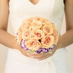 This image captures a quiet, intimate moment before a wedding in Queensbury, focusing closely on a bride's midsection as she holds a carefully crafted bouquet just below her waist. Her wedding dress is a soft ivory, with a lace-trimmed bodice and a gentle V-neckline that hints at traditional elegance, while the skirt falls in smooth, lightly textured folds that frame the flowers. Nestled in her hands is a round, hand-tied bouquet built from dozens of peach and blush pink roses, each rosebud plump and tightly curled, with colours shifting from creamy apricot at the outer petals to a warmer, coral glow at the centre. The arrangement forms a compact dome, like a small glowing sunset cradled against the gown. Encircling the base of these roses is a ring of ruffled blooms in rich purple, their edges softly fading into white, likely lisianthus, adding a cool contrast and a sense of layered depth. Fine sprays of tiny white filler flowers-similar to baby's breath-are woven delicately around the underside, softening the transition between the roses and the bride's fingers. Her nails are neatly manicured in a pale neutral tone, with one thumb showing a glint of subtle glitter, suggesting personal style beneath the formality. The background is blurred and pale, drawing attention to the bouquet and the texture of the dress, as if the bride is standing near a bright window in a Queensbury registry office or a local church hall. The scene feels calm, romantic, and thoughtfully composed, like a bouquet prepared by an experienced neighbourhood florist for a ceremony just moments away.