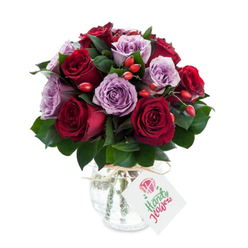 This expertly arranged bouquet pairs dramatic, deep red roses with gentle lavender counterparts to create a balanced, romantic tableau. The crimson and burgundy roses are plush and fully open, their layered petals forming soft ruffles that invite a fingertip to trace their cool, satiny surfaces. Interleaved among them are soft purple and lilac roses whose paler tones provide a calm bridge between the darker reds, producing an overall harmony that is both passionate and soothing. Rich, dark green foliage-likely Ruscus-threads through the arrangement, adding depth and a fresh, leafy scent that complements the floral perfume. Small clusters of bright orange-red rose hips punctuate the massed blooms, offering a cheerful, rustic texture that contrasts with the roses' smoothness. The flowers rest in a rounded, mercury-effect glass vase: antique silvered and slightly crackled, it shimmers with reflected light and gives a subtle vintage note to the composition. Natural raffia is tied around the vase's neck and a plain white rectangular tag hangs forward, bearing a delicate rose sketch and room for a personal message. Photographed against a clean white background, every tone reads vivid and fresh - the deep reds resonate, the lavenders glow, and the greens appear jewel-like. As a local florist would say, this is the kind of bouquet we deliver with care throughout Queensbury, close to Queensbury Station and Honeypot Lane, for birthday surprises, thoughtful thank-yous, or a quietly luxurious home accent.