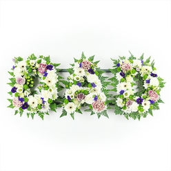 A dignified floral tribute spelling out DAD stretches horizontally across a clean, white backdrop, each letter formed from a richly packed tapestry of fresh blooms and foliage. The main body of each capital letter is built from immaculate white chrysanthemums and daisy-like gerberas, tightly arranged so that no gaps show, creating a smooth, velvety surface that immediately draws the eye. Between these white flowers, a Queensbury florist has woven soft touches of colour: pale mauve and lavender roses, lilac-toned carnations, and the occasional bud poised to open, all adding gentle romance and emotional warmth. Deeper purple blossoms, reminiscent of lisianthus or small pansy-style flowers, are positioned with restraint, their velvety petals bringing a sense of depth, dignity, and reverence that suits a farewell gathering. Around the edges and tucked through the letters run trails of fresh green, including slender fern fronds and glossy foliage, with bright green button chrysanthemums or berries giving subtle texture and life. This verdant outline crisply defines the shape of the D-A-D lettering, just as clearly as a signboard might, yet with far more tenderness. The neutral white background and even lighting ensure every detail is visible, from the ruffled edges of each carnation to the slightly domed centres of the chrysanthemums. The overall effect is of a thoughtful, bespoke sympathy tribute that might be placed beside a coffin in a Queensbury chapel or carried gently to a family grave, expressing enduring love and heartfelt remembrance for a cherished father.