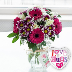 A lavish mixed bouquet glows in bright, diffused light, like it has just arrived with a same-day flower delivery to a Queensbury doorstep. At the heart of the arrangement, generous gerbera daisies in intense magenta and fuchsia command attention, their dark central discs framed by layers of velvety petals that suggest joyful energy. Nestled around them, finely detailed chrysanthemums show off two-toned petals, deep purple melting into pure white at the tips, each bloom centred with a vivid yellow eye that pops against the cooler tones. Soft, creamy white roses are tucked between these bolder flowers, their tightly layered petals adding a touch of romance and refinement. Delicate sprigs of white baby's breath weave gently throughout, creating a lace-like halo that softens the whole design and helps give it a rounded, full profile. Bright green leaves and foliage tuck in behind and beneath the blossoms, giving the impression of something freshly gathered from a well-loved garden near Queensbury Park. The flowers are arranged in a stout, clear glass vase, where you can see the carefully cut stems and the clean water cradling them. Around the neck of the vase, a simple loop of natural twine is tied in a casual bow, from which hangs a small white gift tag decorated with a stylised pink rose and friendly green writing that hints at a handcrafted touch. To the right, a polished, heart-shaped foil balloon catches the light, its silver edge framing a white centre printed with mosaic-style, multi-coloured letters spelling "Love n' Hugs 24-7," tiny hearts sprinkled around the words. The arrangement sits on a pale surface, with a softly blurred, bright background that evokes a peaceful, sunlit room in Queensbury, making this pairing ideal for marking birthdays, anniversaries, or any moment that calls for warmth and affection.