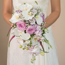 Emanating romance and quiet celebration, this Queensbury bridal bouquet is a lush, cascading arrangement designed to be held close on a wedding morning. The bouquet's focal point is a cluster of pristine white Phalaenopsis orchids, their silk-like petals arching delicately and capturing light in soft, gleaming tones. Around them, full-blown blush pink and lavender roses nestle in gentle harmony, enhanced by the feathery clouds of white baby's breath that evoke an airy sense of magic. Long, magenta spikes of Amaranthus tumble downward, introducing a whimsical texture and movement-echoing the carefree spirit of nearby Roe Green Park. Accents of soft purple stock and unopened buds weave through, supported by glimmers of fresh green foliage that add vigor and a sense of renewal. The bride's lace-embellished gown, glimpsed behind the blooms, speaks to classic elegance and local traditions. The image is softly lit, focusing all attention on each nuance of colour, petal, and curve. Crafted by local Queensbury florists, this bouquet feels both grounded and ethereal, much like the hopes we hold for vows exchanged among loved ones. Each flower's color and scent intertwine, creating a sensory tapestry that brings to mind cherished memories of gardens, joyful celebrations, and refined beauty-perfect for a modern Queensbury wedding, or any romantic day marked by personal meaning.
