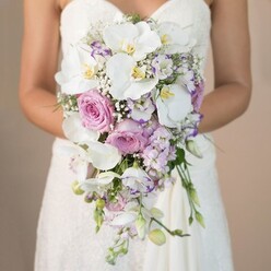 Nestled in the heart of Queensbury, this photograph evokes the artistry of local weddings-a bride seen waist-up holds a breathtaking cascading bouquet, its colours softly glowing against a neutral, light beige backdrop. The floral masterpiece flows in a graceful teardrop, where pure white Phalaenopsis orchids, each with a golden yellow centre, sit alongside lavish, petal-rich blush-pink garden roses. Interwoven throughout the bouquet, sprigs of lavender stock and light purple flowers contribute texture and dimension, while airy clusters of white gypsophila offer a delicate, cloudlike fill. Ruffled lisianthus, edged in deeper purples, peek through, adding a sophisticated touch. Against her strapless white gown, detailed with intricate lace visible on the bodice, the bouquet seems to shimmer-echoing the gentle romance often found along the leafy avenues of Queensbury and its vibrant wedding scene. The subtle scent of roses and fresh greenery, imagined here, would evoke early spring mornings in the nearby parks. As the bride's arms draw in the bouquet, the arrangement's expertly crafted silhouette radiates both elegance and heartfelt celebration-capturing the poetic spirit of a Queensbury wedding and the thoughtful touch of local florists.