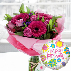 A joyful birthday bouquet and balloon arrangement is styled as though ready to be delivered to a cosy home in Queensbury, perhaps just off Church Lane, radiating warmth and celebration. At the centre, a generous glass cylinder vase holds a lush arrangement of hand-selected blooms, their stems visible through the clear water, suggesting freshness and expert conditioning. Rich fuchsia and magenta Gerbera daisies, with velvety, near-black centres, stretch their petals wide, forming striking focal points that catch the soft indoor light. Nestled between them are layers of blush pink and deeper rose-pink roses, some still tightly furled, others gently opening to reveal spiralling, satin-soft petals that hint at a light, sweet fragrance. Threaded among the main blooms are clusters of tiny, deep purple filler flowers, likely statice, their papery texture adding contrast against smooth rose petals. The design is framed by abundant green foliage, leaves in varying sizes and shapes creating depth, volume, and a natural, garden-picked feel that a local florist from Flower Delivery Queensbury would be proud of. Around the base of the stems, pale pink cellophane or tissue is wrapped and loosely gathered, forming a translucent collar that softens the transition between vase and flowers. Tied around the vase neck with rustic twine, an off-white rectangular gift tag bearing a subtle rose motif hints at a luxury touch without overpowering the design. In the foreground on the right, a round foil Happy Birthday balloon gleams, its white, lightly speckled background dotted with cheerful stylised flowers and circular accents in bright yellow, blue, lime green, pink, and purple. The centre of the balloon proclaims "happy birthday" in sweeping pink script. A softly blurred indoor backdrop, likely lit by daylight streaming from a nearby Queensbury window, keeps focus firmly on this joyful birthday gift ensemble.
