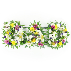 A thoughtful "DAD" floral tribute is shown in full view against a crisp white backdrop, the kind of carefully balanced memorial design often chosen by families in Queensbury saying farewell to a cherished father. Three sturdy letters - D, A, and D - are aligned horizontally, joined subtly underneath by slender, dark green bars so they appear as one flowing word. Each letter is built on a generous cushion of glossy green fern foliage, the feathery leaves softening the outline and echoing the look of a peaceful woodland edge. Packed closely within this frame are masses of fresh, white chrysanthemums and open-faced white gerbera daisies, their petals layered and slightly ruffled, symbolising purity and quiet respect. Scattered through the white are small bursts of sunshine-yellow gerbera daisies and yellow spray roses, which bring a gentle uplift, like a ray of light breaking through a grey sky over nearby Queensbury Park. Interwoven among these are sophisticated purple lisianthus blooms, rich magenta and deep pink spray roses, and tiny lavender-toned flowers, alongside bright green button chrysanthemums that lend a modern, textural accent. The colours blend harmoniously rather than shouting, suggesting warmth, remembrance, and enduring affection. The lighting is even and bright, ensuring that every petal, leaf, and curve of the letters is clearly defined, much as it would appear when placed beside a coffin or photo at a funeral service or wake. This tribute feels both traditional and personal, a bespoke arrangement that could be ordered for same-day delivery across Queensbury and the surrounding streets, offering a heartfelt final message: "Dad, you are loved and remembered."