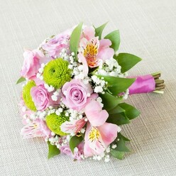 Echoing the spirit of a Queensbury spring and the gentle elegance often found at wedding venues near Cannons Park, this hand-tied bridesmaid bouquet is a masterwork of pastel hues and fresh textures. Roses in varying shades of pink-some blushed with peach, others leaning toward the softest lilac-form the centerpiece of the arrangement, their petals curling in classic spirals that hint at subtle fragrance. Intertwined among them, bright lime button chrysanthemums punctuate the display with their pronounced green and uniquely tactile pom-pom shapes, providing energy and balance. Delicate pink alstroemeria bring an airy softness, their petals adorned with tiny yellow and dark freckles that catch the light like morning dew, nodding to the Peruvian origins of this graceful bloom. Baby's breath (gypsophila) floats throughout in cloud-like sprays, softening lines and imparting an ethereal charm, reminiscent of the early mist that sometimes settles over Queensbury's Lawns Park. Each branch of greenery, including glossy rose leaves and fine-textured filler foliage, provides structure and freshness-binding the colours and shapes into a lush, coherent whole. The bouquet is finished with a radiant fuchsia satin ribbon, carefully tied and leaving the stems visible, reinforcing that genuine, garden-picked feel unique to local floral artisans. Crafted for joyful moments, anniversaries, and the romance of local weddings, this bouquet sings of new beginnings and heartfelt celebration-an artful blend that reflects both Queensbury's natural vibrancy and the attentive expertise of local florists.
