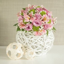 Imagine stepping into a sunlit Queensbury living room, where this elegant floral creation instantly draws the eye. Resting on a crisp white surface, the bouquet bursts with luxe pastel pink and lavender roses, their petals plush and tactile, complemented by playful lime green button chrysanthemums. The gentle curve of the sturdy white woven vase-its intricate open design crafted from slender strips-brings a touch of contemporary flair, reminiscent of artful local finds from Queensbury's markets. Pink alstroemeria, with their striped petals and soft inner markings, infuse the arrangement with breezy sophistication, balancing joy and grace. Sprigs of baby's breath drift between blooms, adding a cloud-like lightness, while glossy green foliage tucks in depth and natural movement. To the side, two artfully positioned spheres in pale woven material-one larger, one smaller-echo the vase's patterns and speak to creative spirit found in home decor throughout the community, from Kenton Lane to Honeypot Lane. A backdrop of textured, muted beige envelops the scene in soothing tranquility. This arrangement, expertly assembled with fresh flowers by local hands, radiates quiet reassurance and confident joy-perfect for birthdays, weddings, or heartfelt anniversary celebrations across Queensbury, and available for delivery from talented florists attuned to the area's unique style.